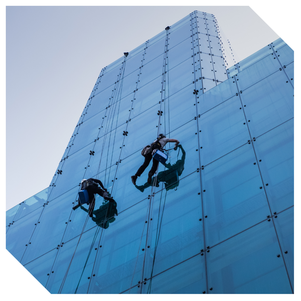 Two workers in safety gear perform exterior window cleaning on the glass facade of a tall, modern building.