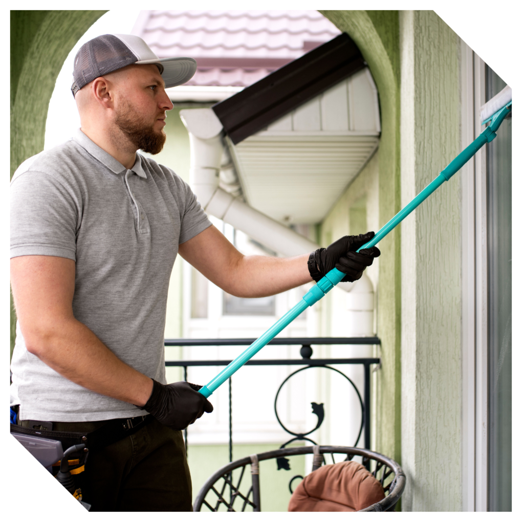 A man in a grey shirt and cap is meticulously cleaning a balcony window with a blue squeegee, donning black gloves for precision—a common sight in the bustling GTA.