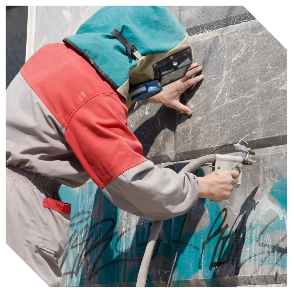 A person in protective gear expertly engages in graffiti removal, sandblasting vibrant paint from a weathered stone wall.
