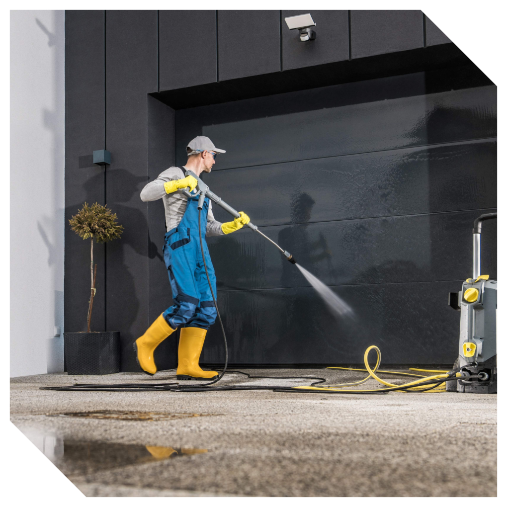 A person in protective clothing expertly pressure washes a concrete driveway next to a modern building, ensuring thorough garage cleaning that leaves the area spotless.
