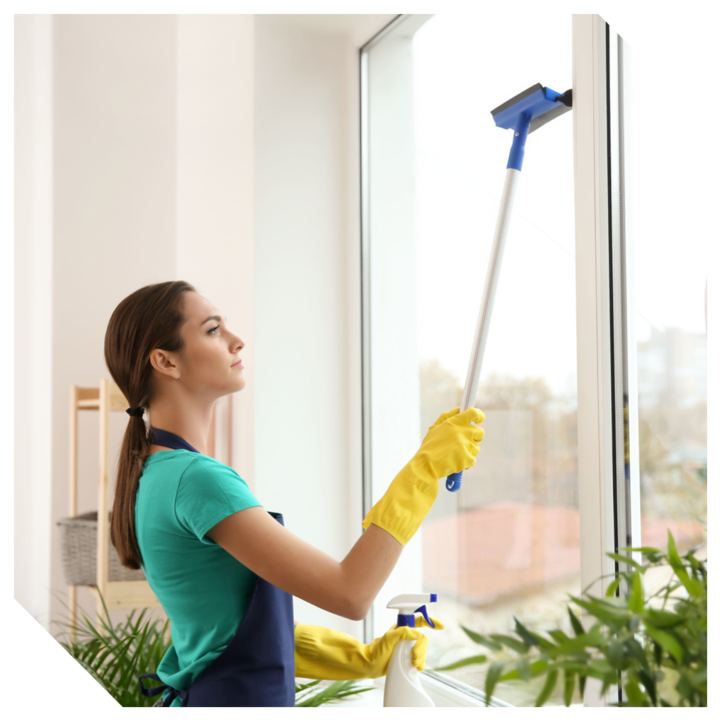 A person in a teal shirt and apron, wearing yellow gloves, is expertly cleaning a large window with a squeegee on the balcony. Beside them are vibrant indoor plants and a spray bottle—a perfect scene for thorough balcony cleaning in the GTA.