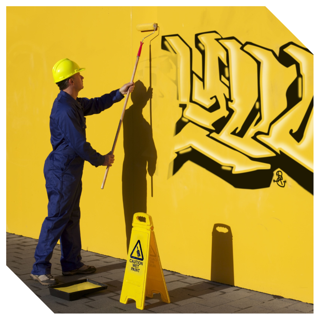 The worker in a blue jumpsuit and yellow hard hat focuses on graffiti removal, carefully painting over the graffiti on the yellow wall with a roller. Nearby, a "Caution Wet Paint" sign ensures safety during the surface restoration process.