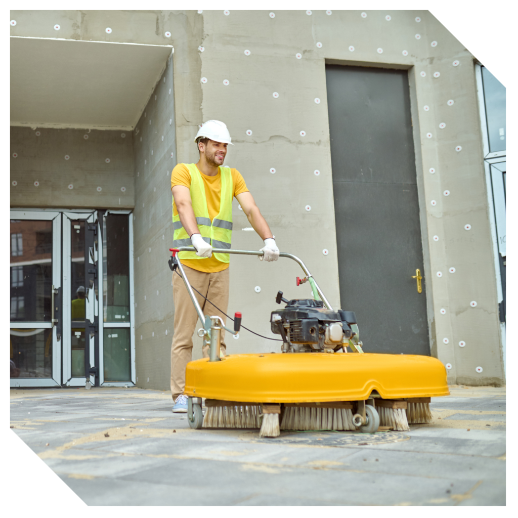 A construction worker skillfully operates a large yellow cleaning machine, ensuring a thorough construction clean up on the paved surface outside the building.
