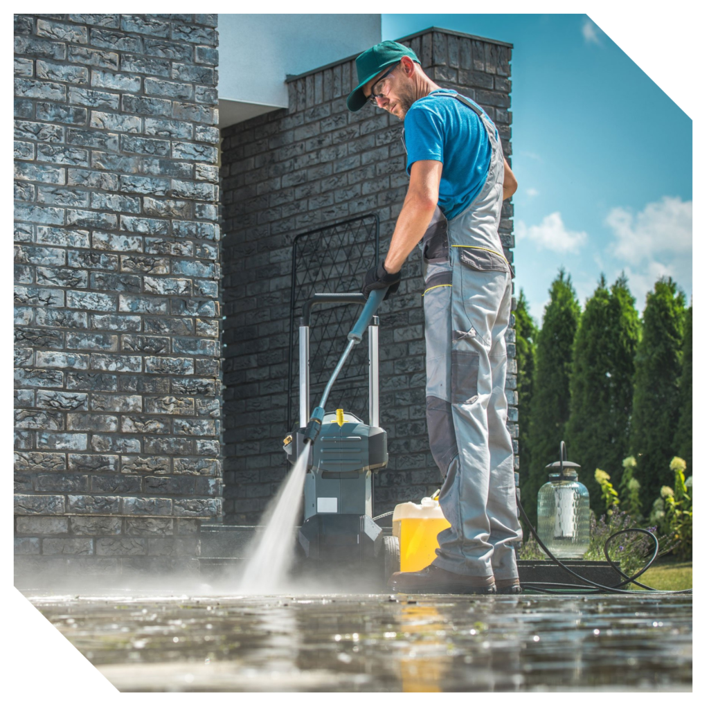 A person in work clothes uses a pressure washer to tackle garage cleaning, meticulously refreshing the paved outdoor area beside a brick building.