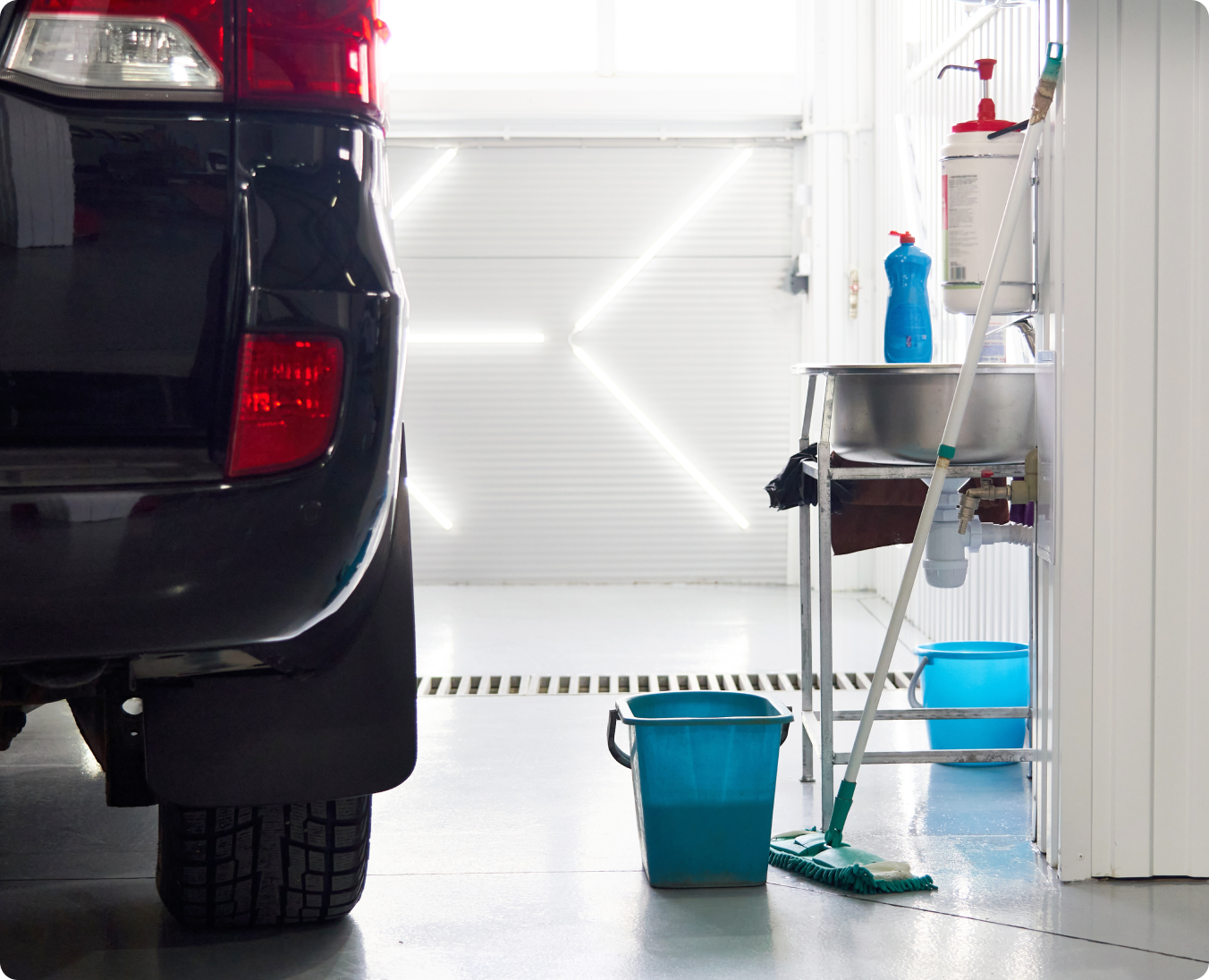 A car is parked in a garage alongside cleaning supplies, including a mop and bucket, next to a sink—a clear setup for an efficient garage washing.